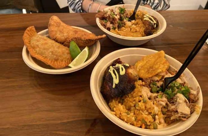 Our-entree-bowls-with-the-roast-pork,-rotisserie-chicken,-rice,-fried-plantains,-and-sauces,-with-a-side-of-the-chicken-and-beef-empanadas Que Chevere ny
