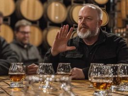 A bearded man gestures while explaining whiskey flavors at a tasting event in a barrel room.